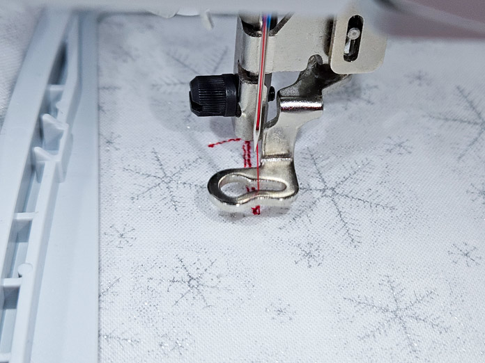 A metal embroidery presser foot with red thread in a sewing machine needle is ready to stitch on white and silver snowflake fabric in a gray embroidery hoop; Husqvarna VIKING Designer Quartz 29, mySewnet Machine Embroidery Software, Husqvarna VIKING Tear-a-Way Light Stabilizer 15 Inches x 25 Yards