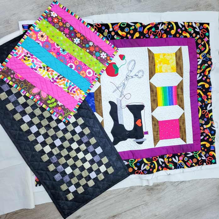A black, gray, and gold table runner, a pink, blue, and brown floral placemat, and a wall hanging with bright sewing-themed appliques