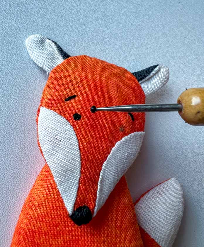 An eye is being added to a small fabric toy fox with a stylus and black paint. PFAFF creative icon 2, PFAFF creative icon 2 sewing and embroidery machine, PFAFF Universal Needles Size 80/12, PFAFF Universal Needles Size 90/14, PFAFF Quilting Needles Size 90/14, PFAFF ¼” Quilting Foot for IDT System, Mettler 40wt polyester thread, oliso iron