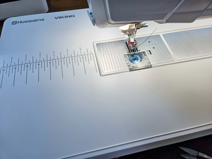 A white Husqvarna VIKING extension table with markings on a sewing machine with a ¼” foot attached. Husqvarna VIKING DESIGNER EPIC 3 sewing and embroidery machine, Husqvarna VIKING 6in/15.2cm Applique Scissor, Husqvarna VIKING Open Toe Presser Foot for IDF System, SULKY Slimline Metallic Dream Assortment