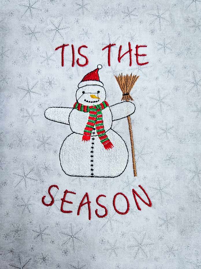 An embroidered snowman on white and silver snowflake fabric with red lettering of tis the season; Husqvarna VIKING Designer Quartz 29, mySewnet Machine Embroidery Software, Husqvarna VIKING Tear-a-Way Light Stabilizer 15 Inches x 25 Yards