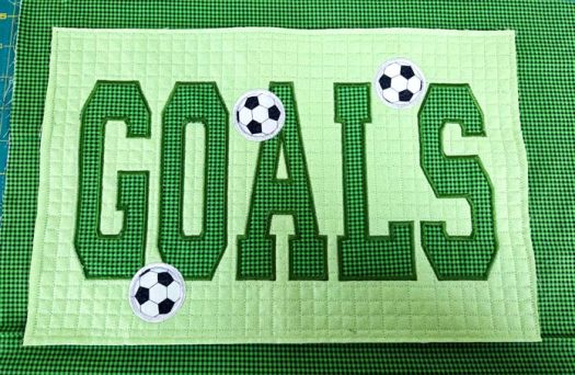 Three fabric soccer balls on green fabric with the word GOALS