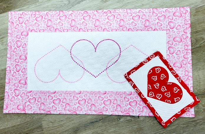 A pink and white table runner with three heart outlines in pink thread and a mug rug with a red applique heart and a border of red fabric