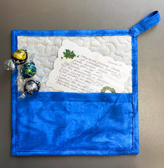 A completed gray and blue quilted potholder with candy and a recipe in the back pocket.