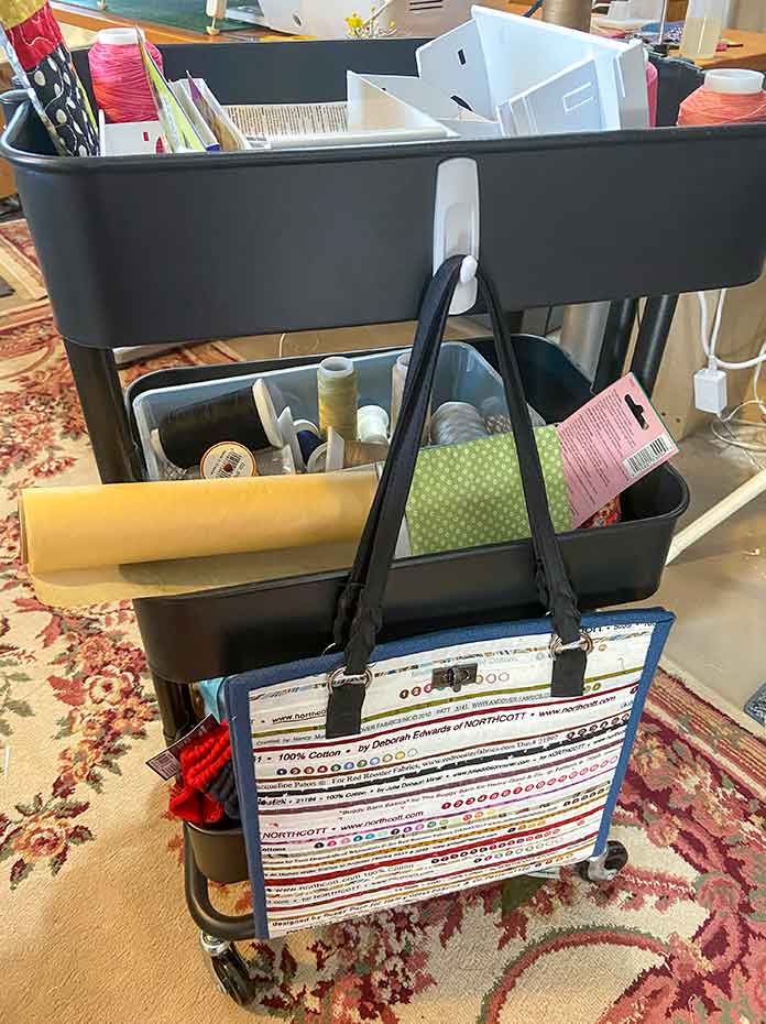  A black 3-tiered rolling cart filled with threads and other sewing items. A bag made of selvages hangs from a hook on the side.