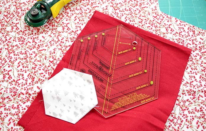 The cutter, winter tree fabric (cut into a hexie), burgandy fabric and berry fabric are shown with the ruler ready to cut hexies, triangles and diamonds. 