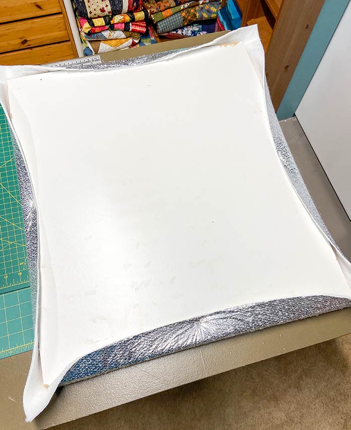 The back of a white wooden board is shown with the shiny Therm Fleece wrapped around it and stapled to the center of each side. UNIQUE Wool Pressing Mat, Oliso M2Pro Mini Project Iron, Oliso Pro TG1600 Smart Iron, UNIQUE Quilting Therm Fleece, Fairfield Toasty Cotton Natural Cotton Quilt Batting
