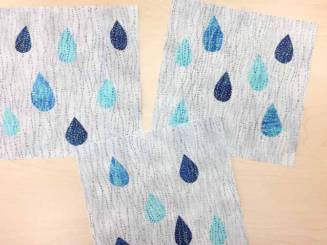 Fabric fused raindrops, in different shades of blue, on three gray quilt blocks. Northcott’s Artisan Spirit Shimmer Echoes.