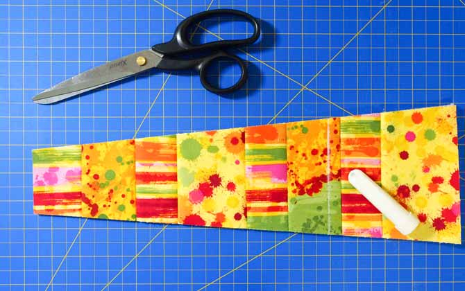 a pair of black handled scissors and multi colored fabric wedge on a blue cutting mat